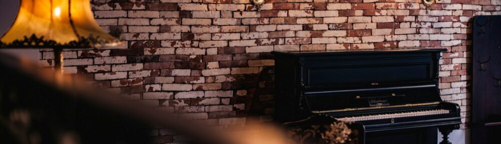 Un pianoforte verticale nero contro un muro di mattoni in una stanza illuminata da appliques, immagine per il teatro in casa