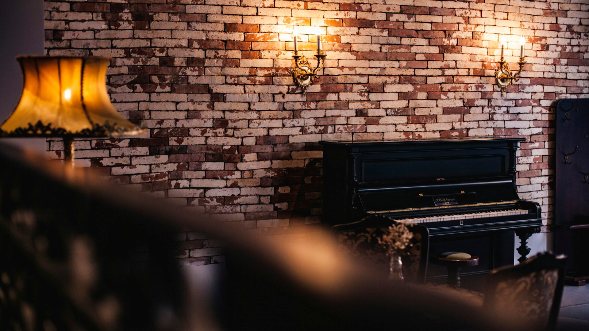 Un pianoforte verticale nero contro un muro di mattoni in una stanza illuminata da appliques, immagine per il teatro in casa