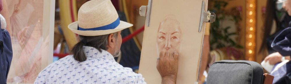 Un ritrattista di strada seduto al cavalletto disegna il volto di una donna, dietro di lui le luci di una giostra