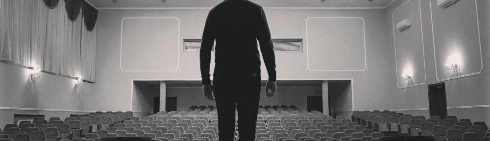 Un uomo solo in piedi sul palco di una produzione artistica guarda le poltrone vuote della platea