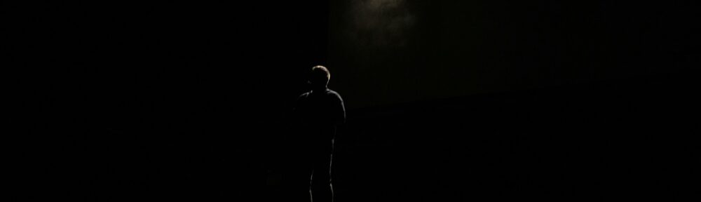 Attore solo e in ombra su un palcoscenico buio, un fascio di luce alle sue spalle