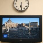 Monitor acceso su Piazza San Pietro in diretta nella reception di un hotel di Roma, con un orologio a lancette sopra