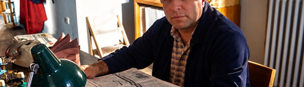 Giuseppe Vitale nei panni del bidello Mario Carrisi seduto alla scrivania con il giornale aperto, immagine per l'auto-promozione degli attori