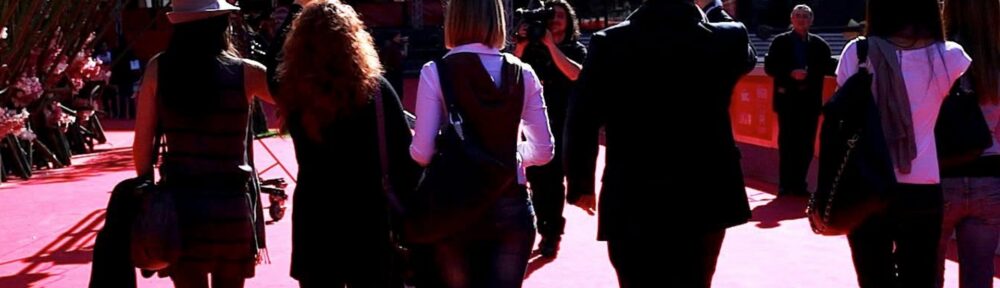 Il red carpet della Festa del cinema di Roma nel 2010, visto da dietro: Giuseppe Vitale in giacca scura saluta con il cast di Come se fosse facile