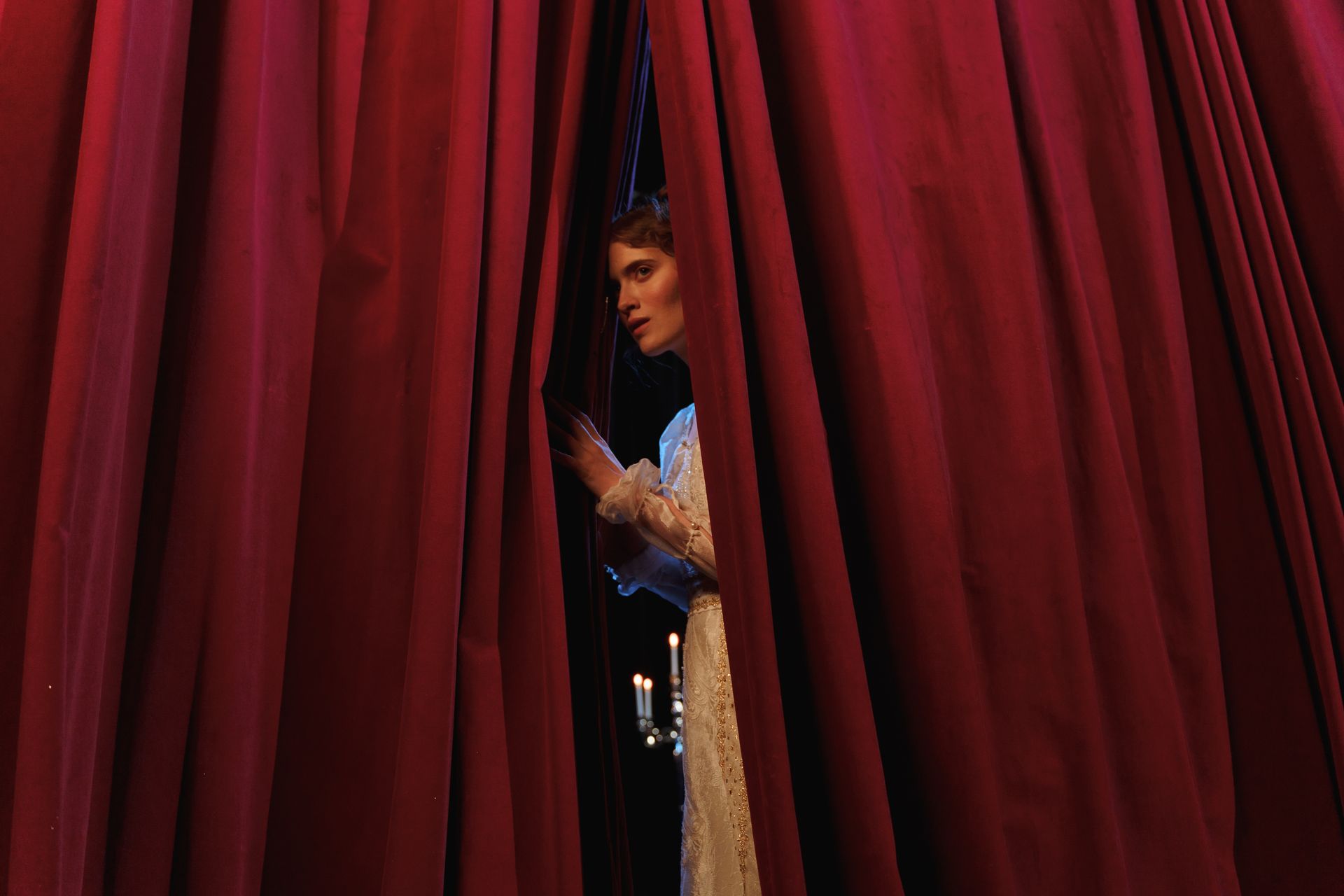 Una donna si affaccia per metà da un sipario di velluto rosso, nascosta dietro le quinte di un teatro